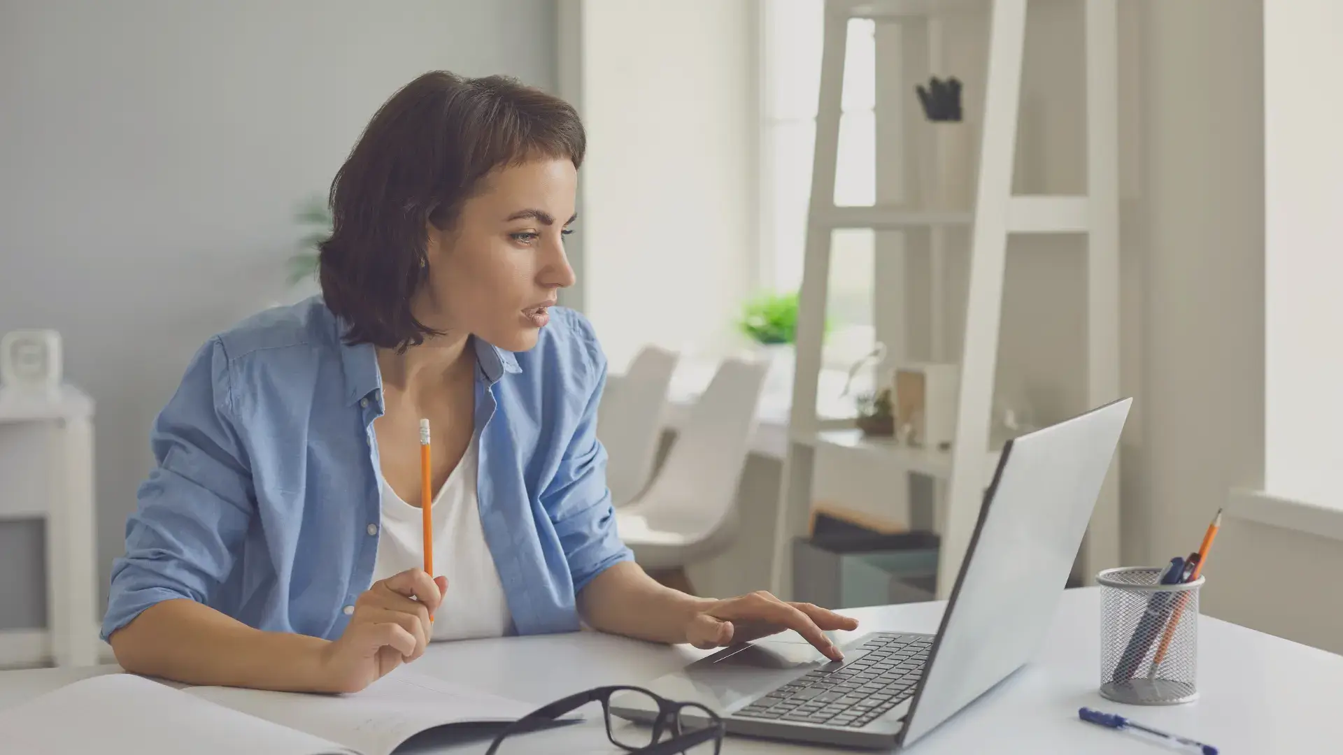 Woman working on a laptop while holding a pencil in a bright office
