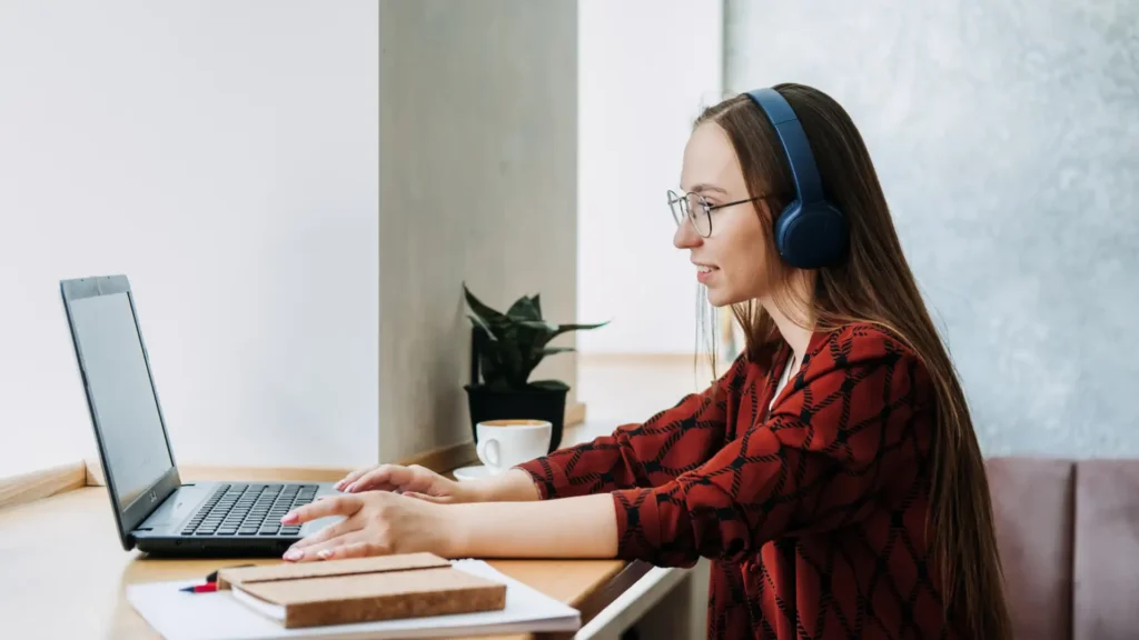 woman with long brown hair, wearing glasses and blue headphones, working on a laptop