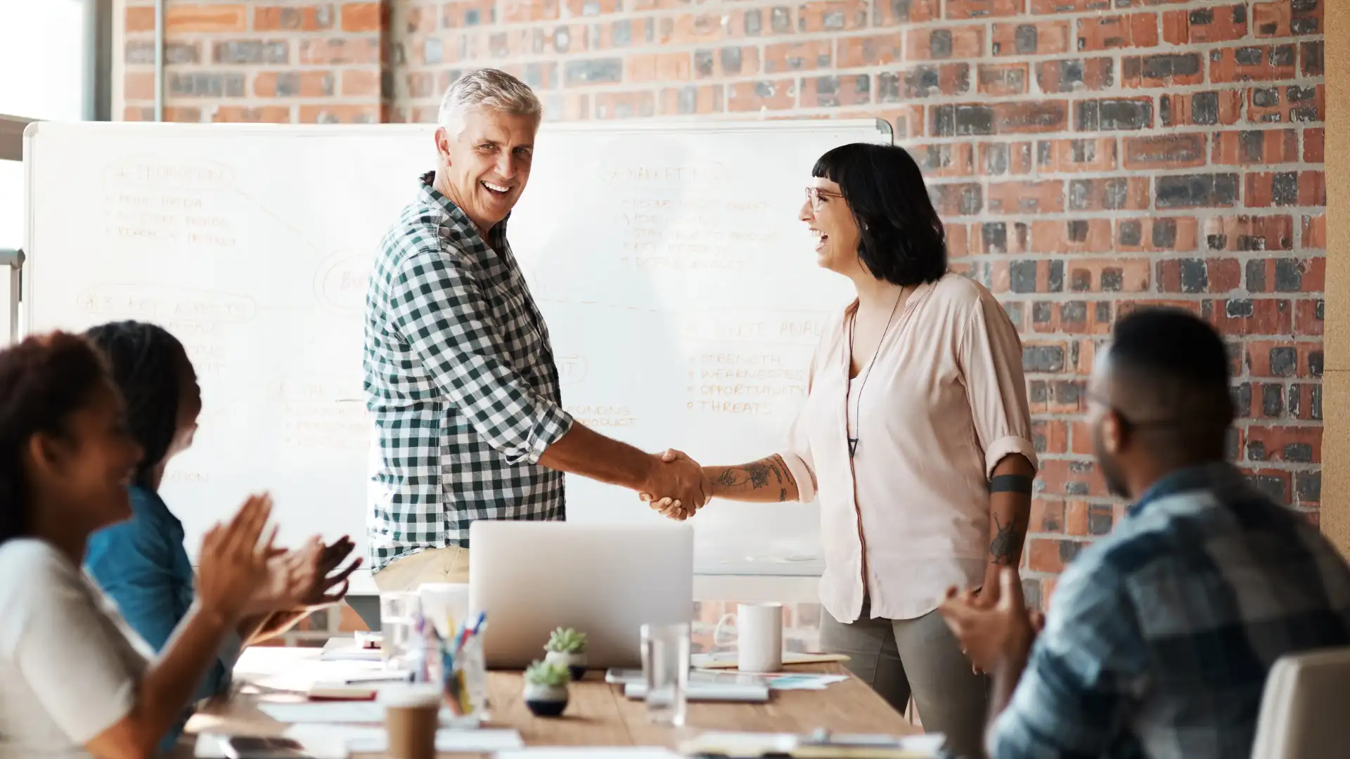 A group of colleagues in a casual office setting applauding as two individuals shake hands in front of a whiteboard