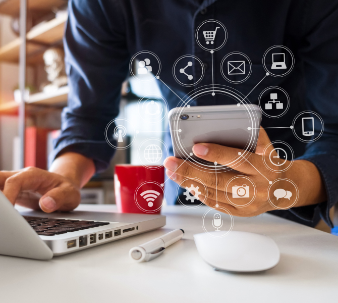 A person using a smartphone and laptop at a desk, with digital icons representing online connectivity, e-commerce, and communication displayed around the phone