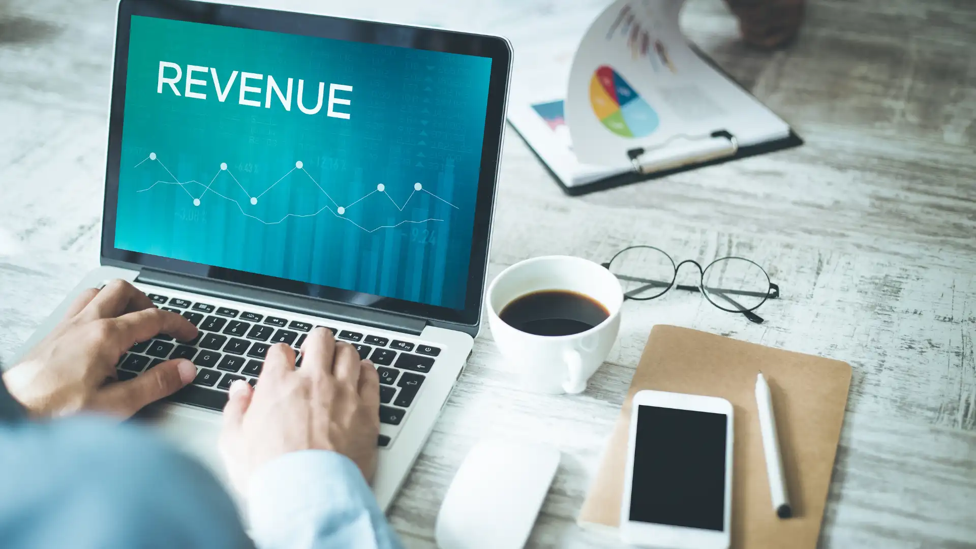 Person working on a laptop displaying "REVENUE" with charts, alongside coffee and documents.