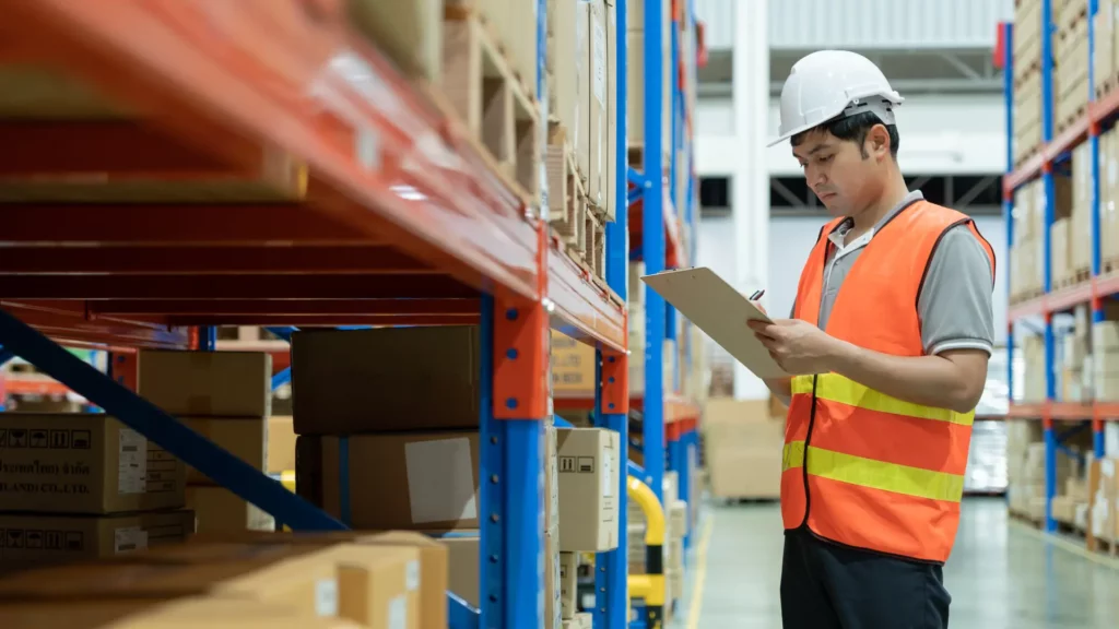 A warehouse worker in a safety vest and helmet checking inventory with a clipboard.