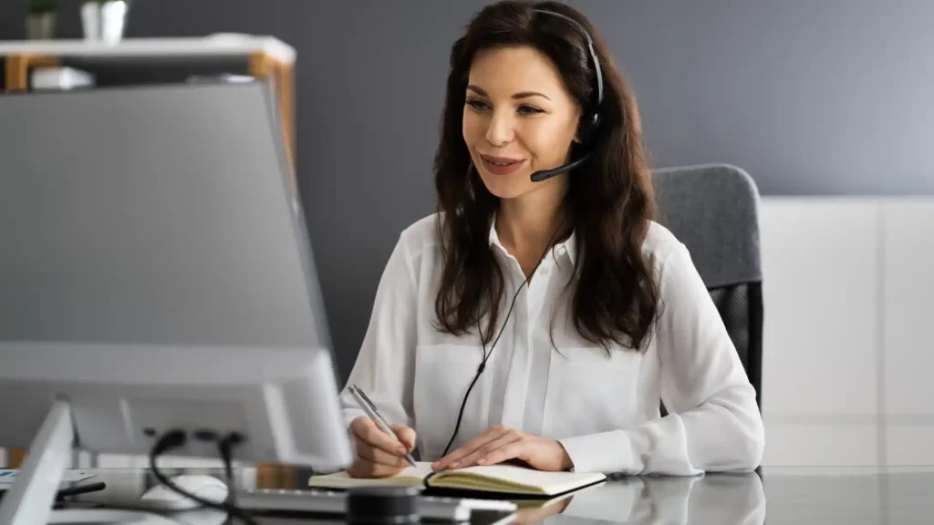 A woman in a headset is smiling while taking notes on a notepad during an online video call at her desk
