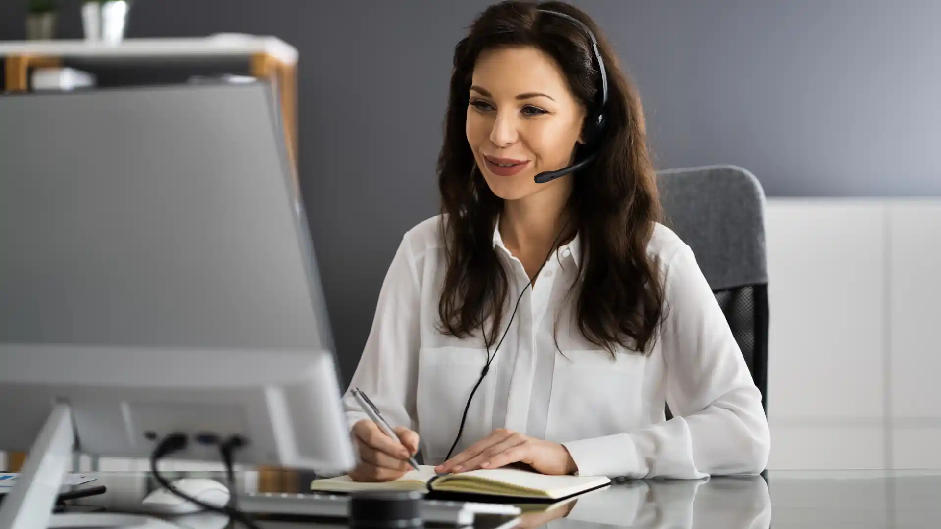 A woman in a headset is smiling while taking notes on a notepad during an online video call at her desk