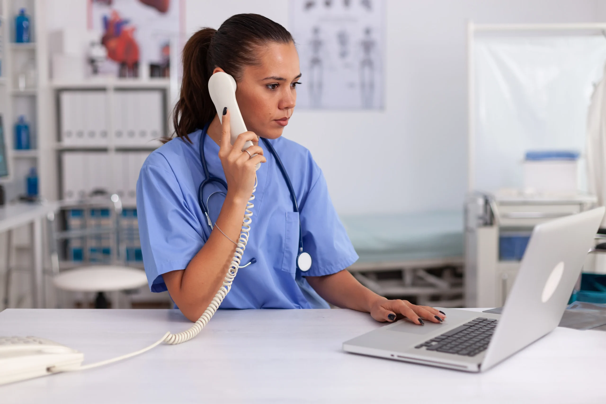 A healthcare worker in blue scrubs speaks on a corded phone while using a laptop at a desk in a medical office.