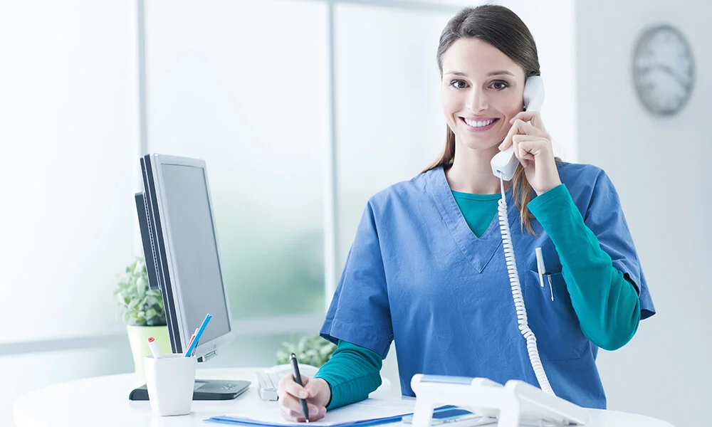 A smiling healthcare worker in blue scrubs speaks on the phone while writing notes at a desk with a computer and office supplies.